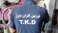 نمای نزدیک لباس کارگاهی با نشان T.K.D روبه‌رو با دستگاه ماشین‌کاری